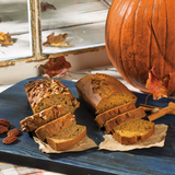 Sliced sweet potato pecan bread and pumpkin bread on a blue surface, with a whole pumpkin and autumn leaves in the background.